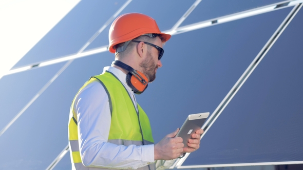 A Professional Is Standing Near a Solar Panel and Working with His Tablet Computer alt