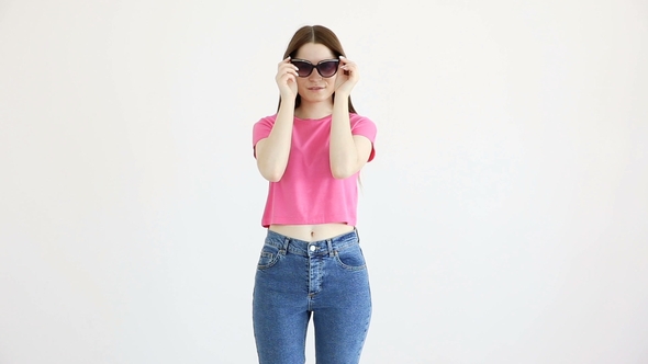 Beautiful Cheerful Girl in Sunglasses, Pink Top and Jeans Posing Against White Wall