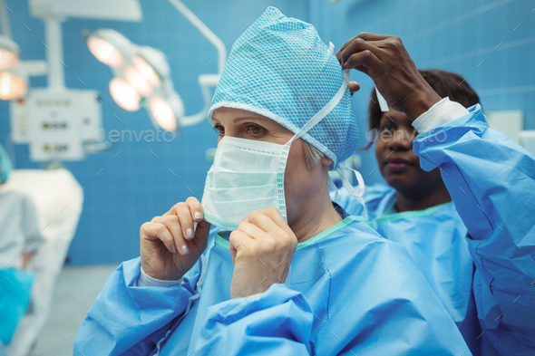 Female surgeon helping her co-worker in wearing surgical mask Stock Photo by Wavebreakmedia