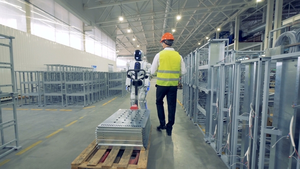 Male Factory Worker Is Launching a Robot Which Starts Pulling a Transporting Trolley alt