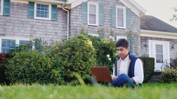 A Young Indian Man Works with a Laptop in His Backyard. Sits on a Green Lawn alt