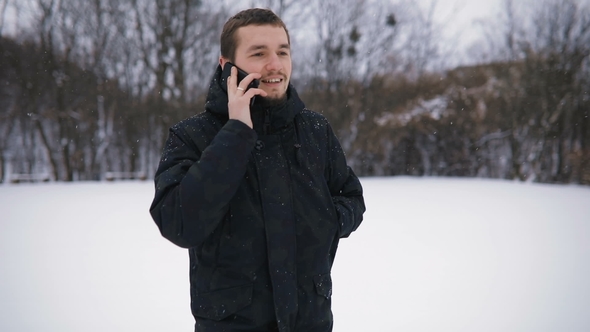 Brunette Young Man Portrait in Black Hood Talking By Phone in Snowy Winter Park