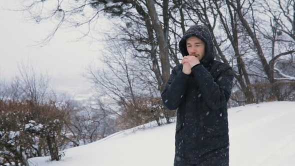 Brunette Young Man in Black Hood Walking in the Snowy Winter Park, Hat Rubs Hands