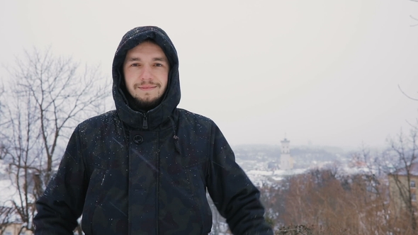 Brunette Young Man Portrait in Black Hood in Winter Snowy Park. . City Town Background View