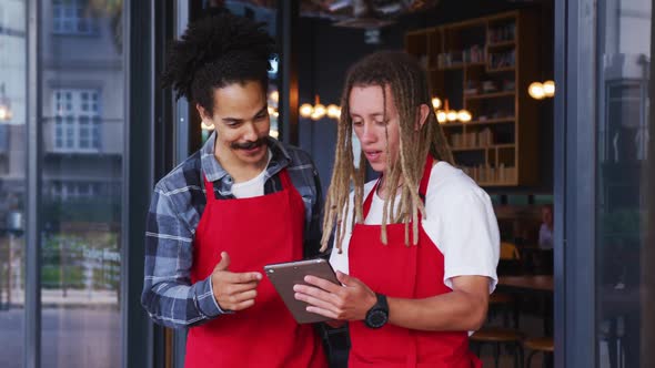 Two diverse male baristas wearing aprons standing in the doorway and using digital tablet alt