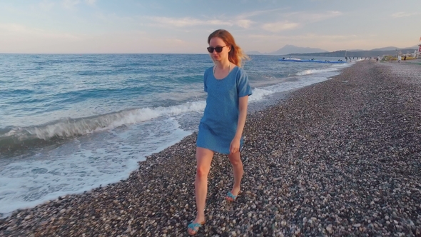 Young Slim Blonde Is Wearing Blue Dress and Sunglasses Is Strolling on Pebble Seashore