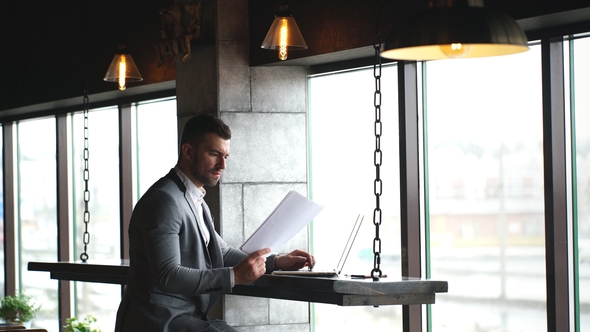 a Young Handsome Businessman Checks Documents and Typing on a Laptop Sitting in a Cafe By the Window