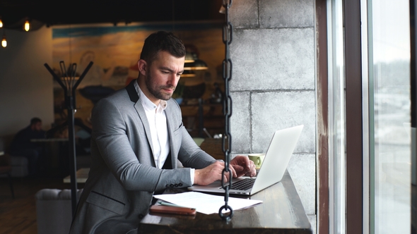 Young Handsome Attractive Bearded Businessman Sitting at the Table in a Suit and Typing on a Laptop