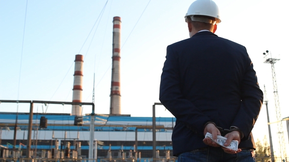 Engineer Chief Stands with Money Dollars in Handcuffs Against the Backdrop of a Power Plant, Rear alt
