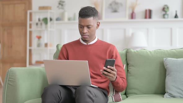African Man with Laptop using Smartphone on Sofa alt
