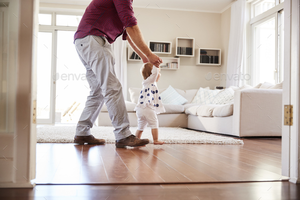 Father helping daughter learn to walk at home, side view Stock Photo by monkeybusiness