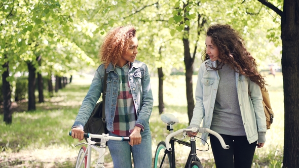 Dolly Shot Of Cheerful Girls Friends Walking In Park With Bikes Dolly Shot Of Cheerful Girls Friends Walking In Park With Bikes
