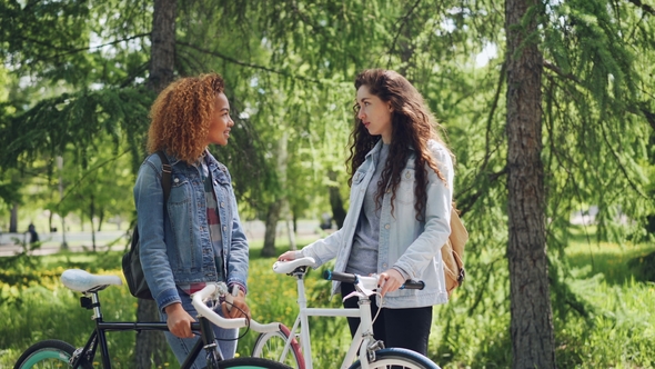 Two Female Bicyclers with Backpacks Are Talking Standing in the Park Holding Bikes alt