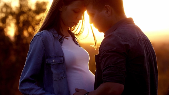Pregnant with Her Partner Hugging Her and Holding Her Belly Outdoors alt