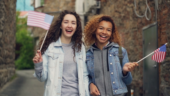 Portrait of Two Pretty Girls in Casual Clothing Waving American Flags and Laughing Looking at alt