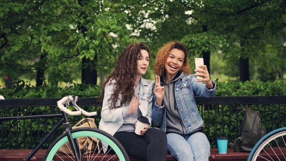 Female Friends Are Taking Selfie with Smartphone Sitting on Bench in Park Holding Coffee, Young alt
