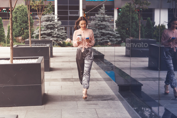 Smiling businesswoman with coffee cup walking on the street and making a phone call Stock Photo by Vladdeep