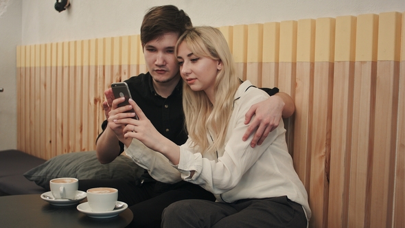 Happy Couple in a Coffee House Having Breakfast and and Using Cell Phone alt