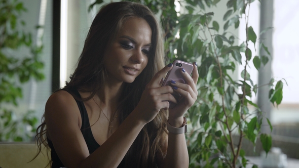 Charming Brunette Girl Is Typing on a Touch Screen of Her Smartphone, Sitting in Restaurant in alt