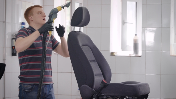 Car Wash Worker Is Cleaning Seat of Automobile in a Room, Using Vacuum Cleaner, Seat Is Standing on alt