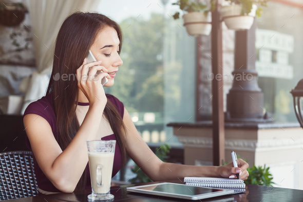 Young businesswoman talking on smartphone and making notes Stock Photo by Prostock-studio