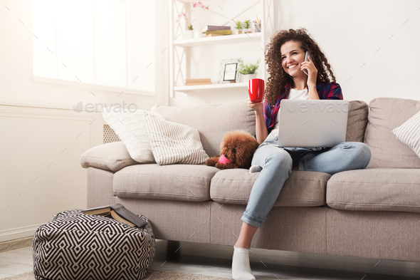 Smiling young woman with mobile, laptop and little dog at home - Stock Photo - Images