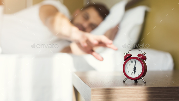 Sleepy guy waking up early after alarm clock signal Stock Photo by Prostock-studio