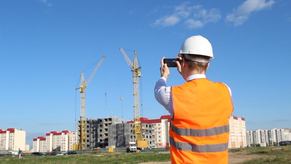 Inspector in a White Helmet and an Alarm Jacket Is Photographing the Construction of an Apartment alt