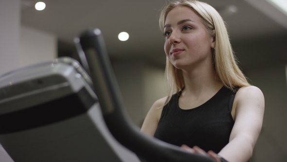Blonde Girl Turns the Pedals on an Exercise Bike Watching the Time in New Gym