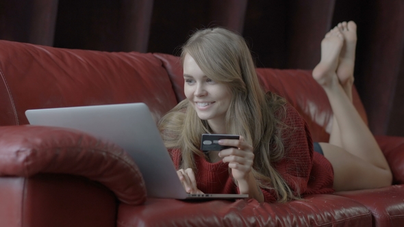 Beautiful Smiling Blonde Woman Leaning on Red Sofa While Using Laptop and Credit Card for Shopping