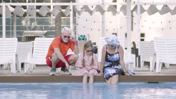 Happy Mature Couple and Their Little Granddaughter Sitting on the Edge of the Pool alt
