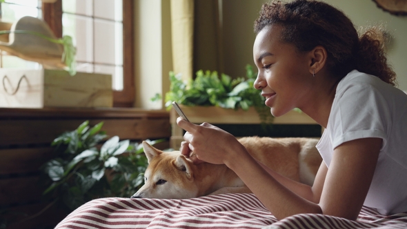 Pretty Girl Student Is Using Smartphone Touching Screen and Laughing Lying on Bed at Home with Cute Dog alt