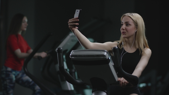 Beautiful Blonde Girl Is Diligent Working on an Exercise Bike and Making Selfie on Her Phone