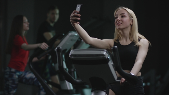 Nice Blonde Girl Is Diligent Working on an Exercise Bike and Making Selfie on Her Phone alt