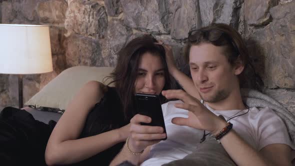 A Young Couple Are Lying In Bed Chatting Looking At Photos On Their Smartphone alt