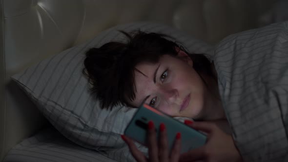 Pretty Brunette Woman Lies in the Bed at Night and Uses the Phone Reading Something and Thinking