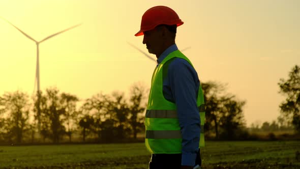 Windmills Rotate Behind Engineer Walking at Substation alt