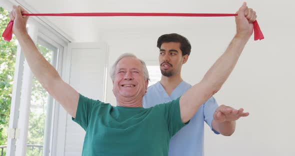 Senior man grimacing while exercising with resistance band against male therapist at nursing home alt