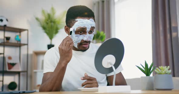 Black-Skinned Guy Looking Into Small mirror when Putting on Moisturiser Mask on face alt