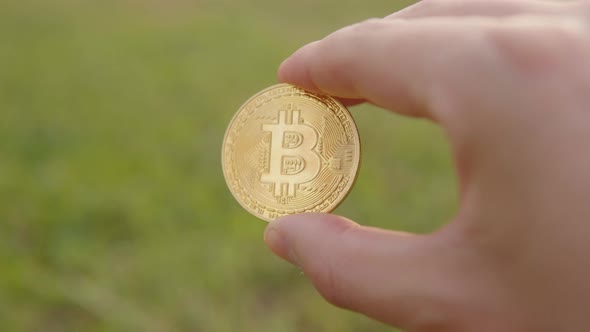 Man Shows a Cryptocurrency Symbol Bitcoin Coin on the Background of a Green Lawn