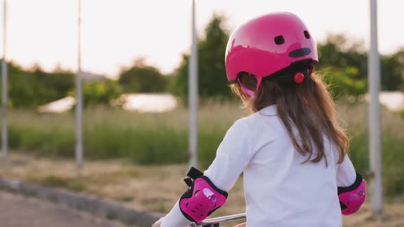 Cute Light Hair Little Girl in Pink Helmet in Elbow and Knee Pads Rides a Bicycle and Turns Back