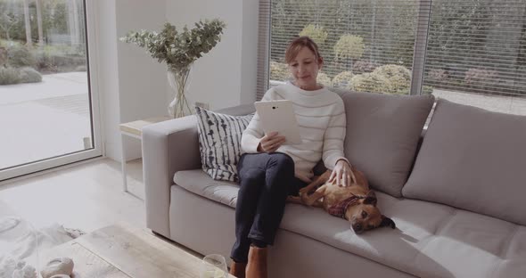 Mature adult female sitting with dog on sofa looking at digital tablet