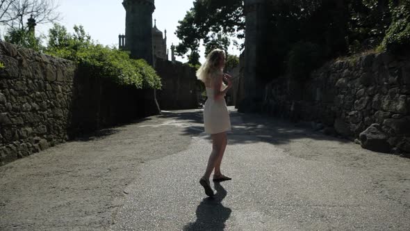 A Young Blonde Girl in a Short Summer Dress Walks on the Road Near an Old Castle