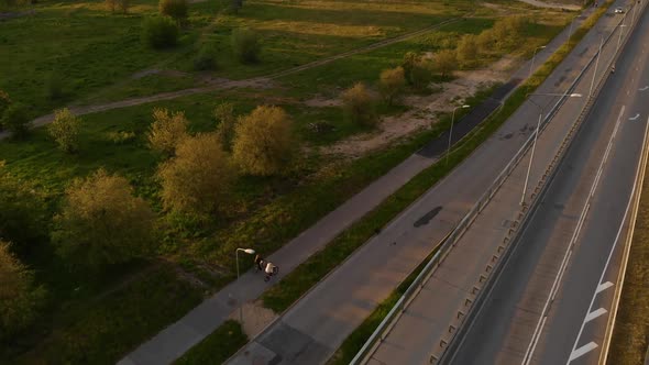 Aerial Backside View: Two People Ride Bicycles Near Highway Bridge During Sunset alt