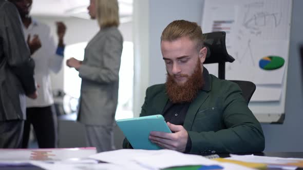 Portrait of Focused Concentrated Caucasian Bearded Man Surfing Internet on Tablet Sitting at Table alt