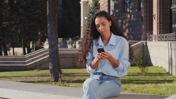 Young Girl Woman Sitting in City Street Looking at Mobile Phone Chatting Online with Smartphone alt
