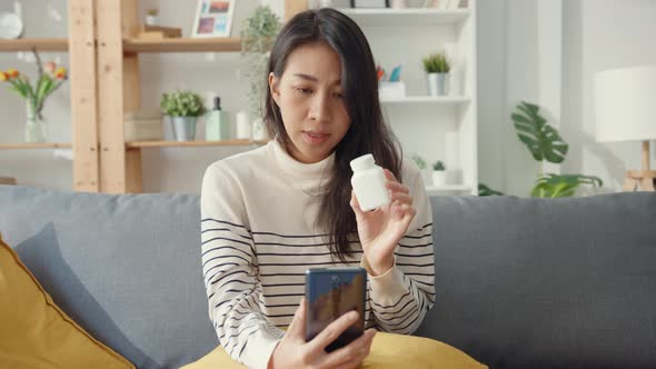 Asian woman hold medicine sit on couch video call with phone consult with doctor at home.