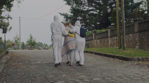 Doctors in protection suits walking with sick woman outdoors alt