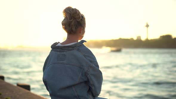 Back View of Young Pretty Blond Girl Sitting on Concrete Pier Enjoying Warm Sunset Shine Looking at alt