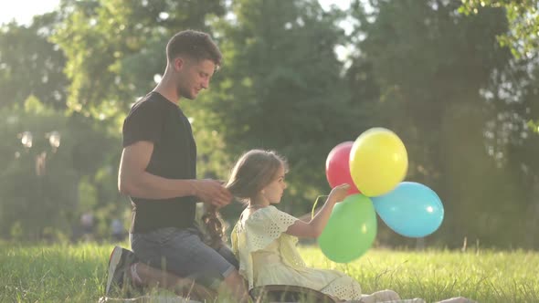 Side View Relaxed Daughter Playing with Colorful Balloons As Young Father Braiding Hair Sitting on alt